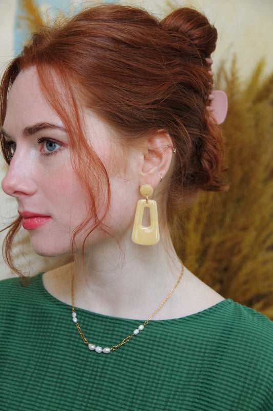 Gorgeous young woman wearing the Audrey earrings in a glossy golden color by the brand Dress To Finesse. This brand is created by Anna De Ceulaer in Antwerp, Belgium. The Audrey earrings are made of clay and are very lightweight. The model is also wearing the Fae necklace which features genuine freshwater pearls.