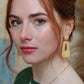 Gorgeous young woman wearing the Audrey earrings in a glossy golden color by the brand Dress To Finesse. This brand is created by Anna De Ceulaer in Antwerp, Belgium. The Audrey earrings are made of clay and are very lightweight. The model is also wearing the Fae necklace which features genuine freshwater pearls.